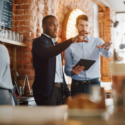 Meeting Behind the Counter and Working on Tablet Computer and Checking Inventory in a Cozy Loft-Style Cafe. Successful Restaurant Managers and Barista at Work