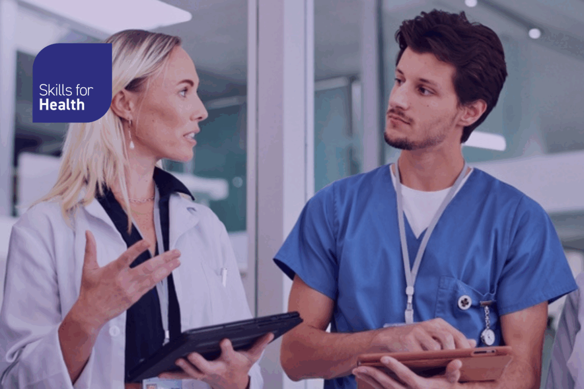 two healthcare staff members talking. one is holding a tablet, they look as though they are discussing a plan of action