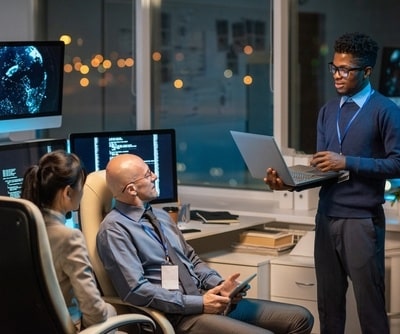 Man stood holding a laptop explaining something to two colleagues