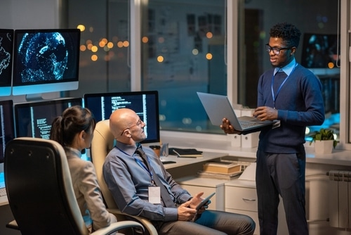 Man stood holding a laptop explaining something to two colleagues