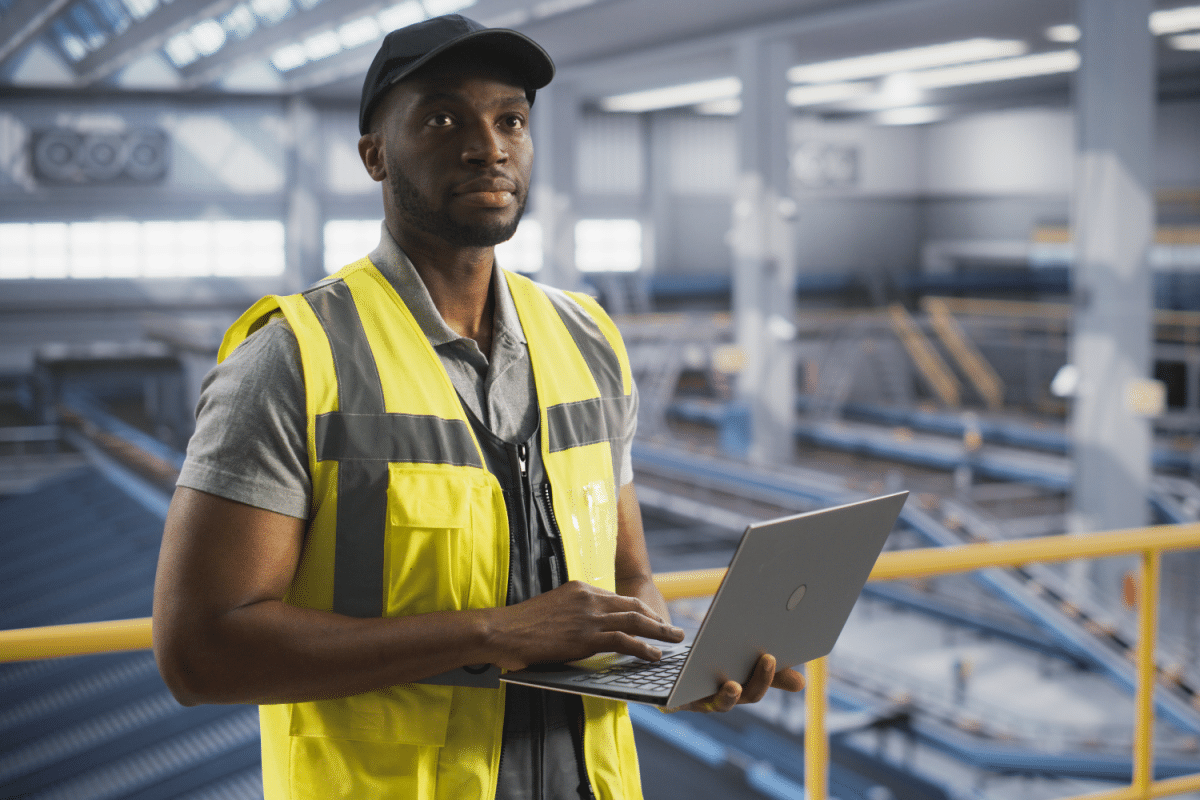 Man wearing a high visibility vest working in a warehouse, holding a laptop